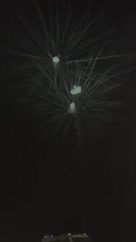 The video captures three small white fireworks exploding in a dark night sky. The fireworks are emitting faint trails of light. In the foreground, the top of a person's head and shoulder are visible on the left, and the top of what appears to be a vehicle or a dark object is visible at the bottom. The overall scene suggests a fireworks display viewed from the ground.