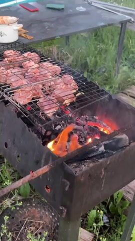 A video displays raw meat cooking on a grill over hot coals. The grill is positioned over a dark metal brazier filled with glowing embers and flames. The meat, appearing to be chicken or pork cuts, is arranged on a wire rack. The background shows a blurred outdoor environment with green grass and what seems to be a table with some items on it. A metal rod is inserted into the brazier, presumably to adjust the coals. The lighting suggests it is either dusk or an overcast day.