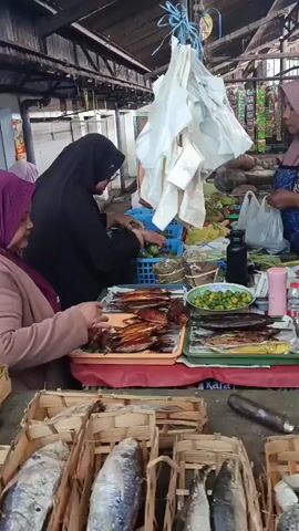 The video shows a bustling outdoor market stall where a vendor is interacting with customers. Several people are present, mostly women wearing headscarves. The stall is laden with various goods, primarily fresh and cooked fish displayed in woven baskets and trays. There are also some fruits and vegetables, as well as bottled beverages and other packaged items. The scene appears to be lively and active, with people engaged in purchasing and selling. The overall environment suggests a typical market setting with a wooden roof providing shelter.