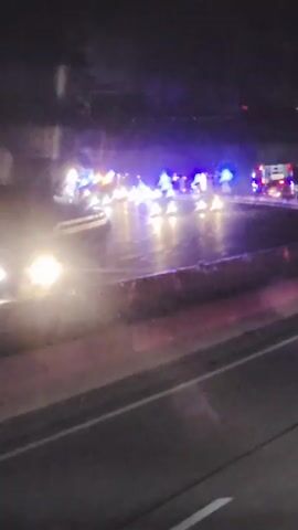 The video shows a nighttime scene on a wet road, possibly a highway. Emergency vehicles, including what appear to be police cars and a fire truck, are present, their blue and red lights flashing. The vehicles are stationary and arranged in a line, suggesting an incident has occurred. The road surface is reflective, indicating recent rain or dampness. The surrounding environment is dark, with trees visible in the background. The perspective suggests the video is being recorded from a moving vehicle.