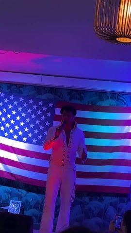The video features a person dressed as Elvis Presley, singing into a microphone on a stage. They are wearing a white jumpsuit with embellishments. Behind them is an American flag, with additional backdrop elements suggesting a tropical theme. The stage is lit with multicolored lighting, creating a lively atmosphere. There are audience members, some holding up their phones to record the performance. A modern lamp fixture is visible hanging from the ceiling.