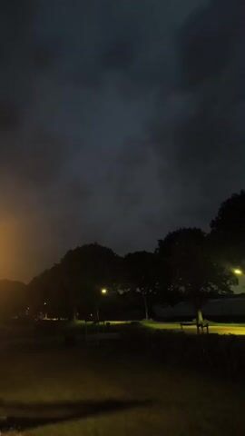 The video displays a dark, cloudy sky with some streetlights illuminating trees and grass. The scene appears to be a park or a residential area at night. The lighting is dim and the colors are muted. There are no people or animals visible in the video. The atmosphere is calm and quiet.