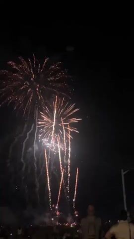 The video shows a fireworks display at night. Several bursts of orange and white fireworks are visible in the dark sky. Below the fireworks, a crowd of people is gathered, silhouetted against the light. Some individuals in the foreground are facing away from the camera, looking towards the fireworks. The environment appears to be an outdoor gathering, possibly a beach or open field, given the dark, flat ground and the presence of many spectators. The fireworks are the main focus of the video, with multiple explosions and trailing smoke adding to the visual spectacle.