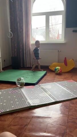 A wide shot shows a toddler in a blue-and-gray striped shirt and khaki shorts standing near a green gymnastic mat. He walks closer, squats, and appears to be watching something out of frame to his right. A gray folding mat lies in the center foreground. In the background are a window, a radiator, a yellow plastic toy, and a multicolored ball. A TV is on the wall. The floor is tiled. The video is lit by natural light from the window.