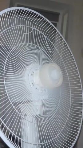 A white electric fan is shown from the front, close-up, with its blades rotating. The fan has a white grill and a white central motor housing with a beige cap. The background is out of focus and appears to be an indoor setting with a wall and a window or door frame visible. The fan appears to be on and in operation, with some wisps of mist or vapor visible around the central part of the fan blades. The video is very short, lasting only a few seconds, and focuses entirely on the rotating fan.