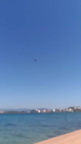 A helicopter flies across a clear blue sky over a body of water. The helicopter is dark in color, with visible rotors and a tail boom. Text reading "POLIS" is visible on the side of the helicopter. In the distance, a coastline with buildings is blurred and indistinct. The scene appears to be a sunny day, likely in the afternoon given the bright sky. The location is identified as Urla, Turkey. The overall impression is one of a clear, open sky and a coastal environment.