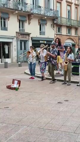 A band of eight musicians plays on a public square in front of buildings. The group includes trombone players, saxophone players, trumpet players, and a drummer. The musicians are casually dressed in patterned shirts, jeans, shorts, and sandals. There is a money collection box open on the ground. The background includes buildings with balconies, awnings, and shops.