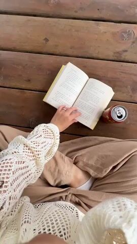 The video begins with an overhead shot of a person's hands holding an open book and a soda can placed on a wooden surface. The camera then pans up to reveal a young woman sitting on a wooden floor inside a wooden structure with ornate railings. She is wearing a white, open-knit sweater and tan pants. She smiles at the camera, holding it out as if taking a selfie, with the open book and soda can visible beside her. The environment appears to be outdoors, possibly a park or garden, with greenery visible in the background.