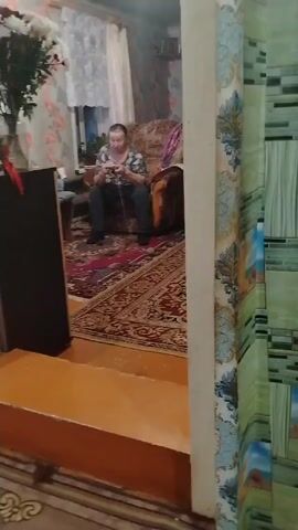 A woman is sitting on an armchair, knitting with yarn. She is wearing a patterned shirt and dark-colored pants and socks. The armchair is patterned and situated on a rug with a red and gold design. Next to the armchair is a small table and a speaker. The room has floral wallpaper and a window with curtains. A ball of yarn sits on the rug near the woman's feet.