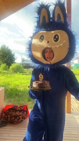 A person in a blue furry monster costume with large ears and a cartoonish face holds a chocolate cake with a lit candle on it. The person is standing on a wooden deck outdoors. A group of young girls are gathered in front of the person, watching. The background shows a green lawn, trees, houses, and a cloudy sky. One of the girls smiles and claps her hands. Another girl in a blue dress stands closer to the person in costume. A third girl in a gray dress stands to the side. A fourth girl in a black shirt and pants is partially visible. A fifth girl in a light blue long-sleeved shirt and purple pants is also visible. There is a large red bag with colorful designs on the left side of the frame. A small blue inflatable pool is visible in the background. The video shows the person in costume presenting the cake to the girls.
