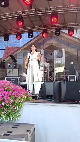 A woman in a white outfit is singing on a stage. She has long dark hair and is holding a microphone. A man is playing the bass guitar to her right. Another man is playing the drums behind them. There are speakers and other musical equipment on the stage. Pink flowers are visible in the foreground. The stage is covered by a roof with red lights.