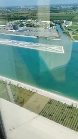 The video pans over a large turquoise body of water with rectangular artificial islands. In the background, there are buildings, palm trees, and manicured lawns. The water appears to be part of a resort or a man-made attraction. The overall scene suggests a modern, well-maintained outdoor space.
