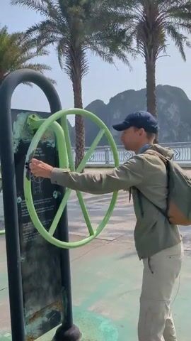 A man in a baseball cap, khaki pants, and an olive green jacket is standing next to an outdoor exercise machine. The machine is a large, light green wheel attached to a black frame, with an informational sign next to it. The man is using both hands to rotate the wheel. He is wearing a backpack. The background includes palm trees, a waterfront railing, and mountains in the distance under a clear sky. The ground has a painted green and white pattern. Someone is cheering him on in Russian.