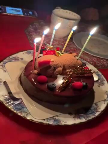 A close-up video shows a chocolate cake on a patterned plate, adorned with two teddy bear figures lying on top, raspberries, blueberries, and golden decorations. Five lit candles are placed around the cake. The background is a red tablecloth, and a smartphone and mug are visible in the periphery. The lighting is dim, creating a warm glow around the candles.