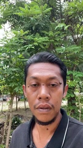 A man in a black shirt stands outside in front of trees. The man is looking directly at the camera. There are banana trees behind him. To the left of the man, there is a building with a yellow wall and a red door. There are also some trees in the background.