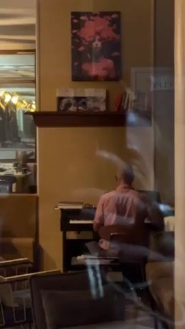 A man wearing a pink collared shirt is playing a keyboard. He is bald and seen from the back. He is seated on a chair in front of the keyboard. Above the keyboard, there is a shelf with some books and framed pictures. A painting of a woman with pink hair is hanging on the wall above the shelf. The scene appears to be indoors, possibly a room with furniture reflected in a window or glass surface in the foreground.