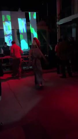 A short video clip shows a dimly lit outdoor space with red and pink lighting. In the foreground, a person in a long gray dress and a white cap is dancing with their arms raised. Behind them, a person in a red shirt and dark pants appears to be near DJ equipment. Another person stands to the right of the dancer. In the background, abstract projections of green and red shapes, and a geometric pattern are displayed on large vertical screens. The overall atmosphere suggests a nighttime event or party.