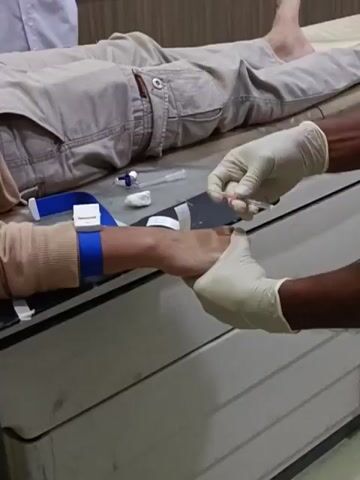 A person lies on a medical examination table, wearing light-colored pants and a long-sleeved shirt. A tourniquet is visible on their left arm, which is resting on a black pad. A person wearing white gloves is holding the arm and inserting a needle into a vein, with a small tube attached to the needle. Another person in a white shirt is partially visible in the background. The room appears to be a medical facility.