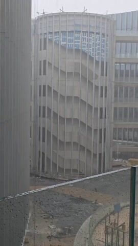 The video shows a cylindrical building in the rain. The building has a geometric pattern on its exterior, and other buildings are visible in the background. Raindrops are visible on a glass surface in the foreground. The overall scene is gray and overcast. An indiscernible voice is heard.