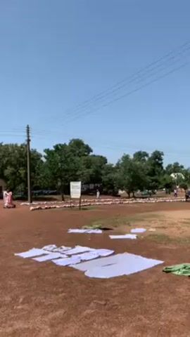 The video depicts an outdoor scene, possibly a courtyard or large open space, on a sunny day. The ground is mostly reddish-brown dirt, interspersed with patches of dry grass. In the foreground, several pieces of white or light-colored cloth are laid out flat on the ground, presumably to dry.

In the mid-ground, there are trees and what appears to be a string or line along which white cloths are hanging. A small, weathered building with a light-colored facade and a dark roof is visible. There are also people walking in the area.

In the background, there is a parking area with a vehicle. Several people wearing dark clothing and white head coverings are present. 

The sky is clear and blue with power lines overhead. Overall, the scene has a rural or semi-rural feel to it.