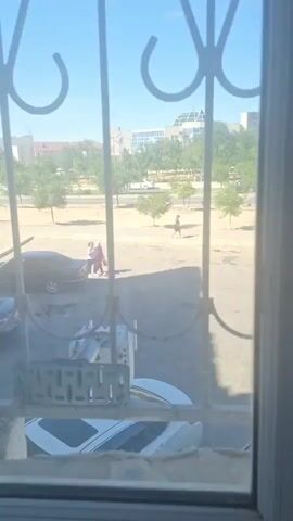The video captures an outdoor scene through a barred window. In the foreground, a white car with a sunroof is partially visible. In the mid-ground, two individuals are standing close together, one wearing a red garment and the other in lighter clothing. To their left, a dark-colored car is parked. Further in the background, a street with trees and some buildings can be seen. Another individual is walking on the sidewalk in the background. The sky is clear and blue.