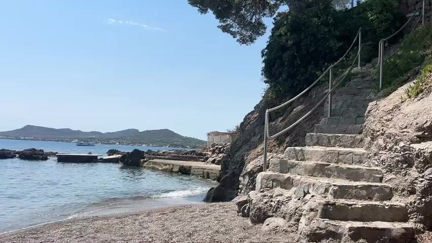 A stone staircase with a metal handrail curves up a rocky incline on a sunny day. The staircase is situated next to a pebble beach and the calm blue ocean. In the distance, a boat is visible on the water. Lush green vegetation and trees grow along the top of the incline.