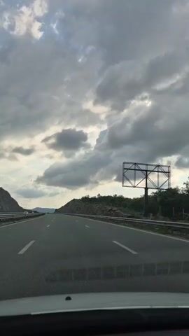 A forward-facing view from inside a moving vehicle displays a multi-lane highway under an overcast sky. The road, marked by white dashed lines, is flanked by metal guardrails and rocky, vegetated hillsides. A green road sign is visible on the right hillside, and a metal billboard structure stands on the far right. The sky is covered with grey clouds, showing some lighter patches. A single white vehicle is distantly visible in the left lane.