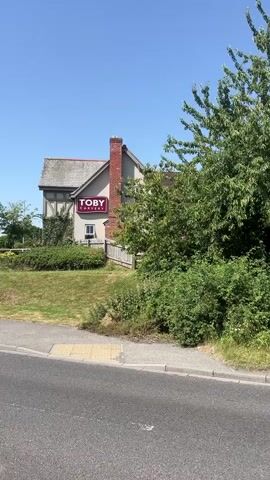 This is a video of a Toby Carvery restaurant viewed from the side of a road on a clear, sunny day. The building is light brown with a grey roof and a prominent red brick chimney. A large red sign with white lettering that says "TOBY CARVERY" is visible on the side of the building. There is a grassy area with some bushes and trees in front of the restaurant, and a paved walkway leading to it. The road in the foreground is asphalt with white lane markings. In the background, there are streetlights and some other buildings visible, along with more trees and landscaping. The sky is a clear blue.