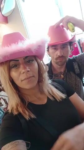A woman with blonde hair and a man stand side-by-side, both wearing pink cowboy hats. The woman's hat has a white feather boa trim. She wears a black top and a black bag across her chest, while the man wears a patterned shirt and a black backpack. Both individuals have tattoos visible. The woman sticks her tongue out, then makes a peace sign towards the camera. The background appears to be a clothing store with various garments hanging on racks.