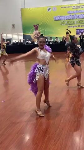 This video shows a dance competition taking place on a polished wooden floor in a large hall. A large banner with Vietnamese text and graphics hangs on the back wall. In the foreground, a young female dancer in a sparkly silver costume with purple feathered accents is performing. She has her hair pulled back and is wearing high-heeled shoes. Her costume includes a short, fringed skirt and a decorative feather boa around her waist. She is captured in mid-motion, with her body angled and her arms extended. To her right, another female dancer in a similar, darker costume is also dancing, and further to the right, a male dancer in a suit is visible, possibly a judge or another participant. In the background, several judges or officials are seated at a long table behind the dancers. The lighting appears to be bright, typical of an indoor sporting or performance venue. The overall scene depicts a competitive dance event.