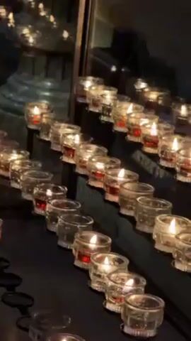 The video shows several rows of small glass candles, with visible red liquid inside, placed on a metal shelf against a reflective background. The candles are lit, casting a warm glow. The background is blurry, suggesting an interior space, possibly a church. The camera is static, focusing on the candles. There are no visible people or interactions in the scene.