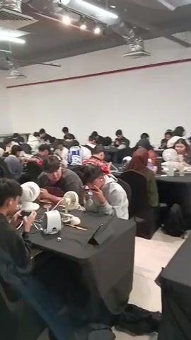 This video shows a large group of people, predominantly young males, gathered indoors and seated at tables covered with black cloths. Most individuals are focused on their mobile phones, with some wearing earphones or headphones. Several tables are equipped with small desk fans and various electronic devices, including what appear to be controllers and charging cables. The environment appears to be a well-lit indoor space with white walls and a visible ceiling structure with lights and conduits. The overall impression is that of a mobile gaming tournament or gathering.