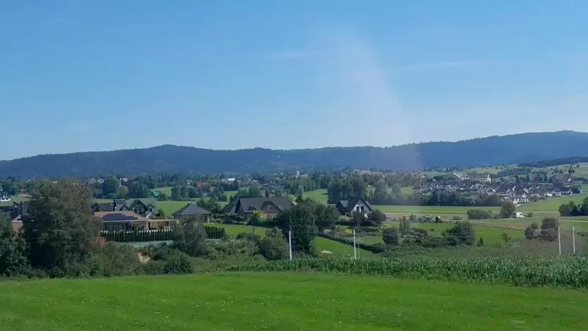 A drone shot reveals a panoramic view of a rural village nestled in a valley. The village is surrounded by rolling green hills and a backdrop of distant mountains under a clear blue sky. The houses are scattered throughout the landscape, featuring a mix of traditional and modern architecture with some exhibiting dark-colored roofs. Trees and lush green fields are interspersed between the buildings and on the slopes of the hills. Some of the houses are quite large and appear to be well-maintained. In the foreground, the landscape is dominated by a vibrant green grassy field. The overall impression is one of a peaceful and picturesque rural setting.