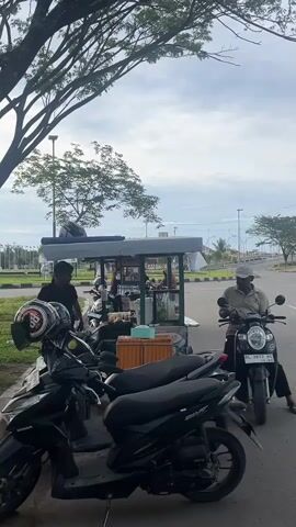 A street food cart is parked on the side of a road under a large tree. Several motorcycles are parked in front of the cart. A man in a dark shirt stands behind the cart, appearing to serve food or prepare items. Another man wearing a hat sits on a black motorcycle to the right of the cart, facing the road. He is looking down at his phone. The sky is cloudy.