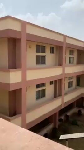 The video shows a three-story building with a courtyard, possibly a school or a dormitory. The building is painted in light yellow with pink trim. The courtyard has a grassy area with some trees. A walkway connects the building to another similar structure. The sky is partly cloudy. No people are visible. The camera pans across the scene.