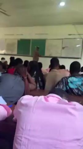 The video shows an interior scene of a classroom filled with students. The camera moves slowly across the room, focusing on the students who are sitting at desks, apparently taking notes or listening to a lecture. Most of the students are facing forward, towards the front of the classroom, where a person is standing in front of a chalkboard. The person at the front is wearing a patterned shirt and appears to be teaching or presenting. The classroom has several windows, a ceiling fan, and a few chalkboards with writing on them. Many of the students closest to the camera wear pink shirts. The overall lighting in the room appears dim.