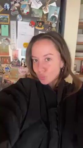 A woman with light brown hair, fair skin, and wearing a black jacket is centered in the frame, looking directly at the camera. She is puckering her lips as if to give a kiss. Behind her is a refrigerator covered in magnets, photos, and notes. To the right is a shelf unit with books and other items. The lighting is bright and appears to be natural.