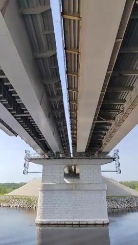 The camera is pointed upwards, showing the underside of a bridge from the water. The bridge has two separate sections, both grey. The support column is made of brick and has a rectangular opening. Small rocks line the water's edge, and there is green vegetation visible in the background. The sky is blue with very few clouds. The camera pans slightly from left to right.