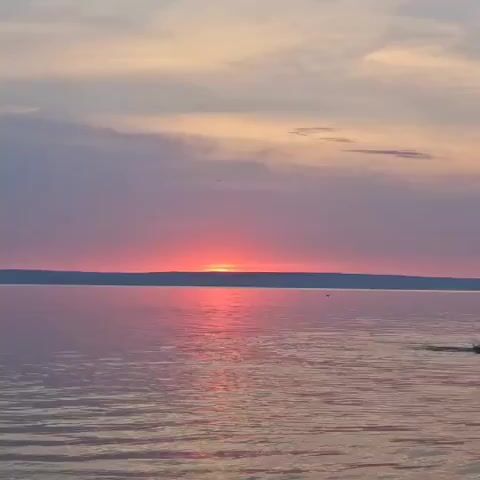 A body of water reflects the colors of a sunset. The sun, a bright orange, is partially obscured by a dark horizon line. The sky above is a blend of pink, purple, and yellow hues, with wispy clouds. Small ripples on the water's surface catch the light.