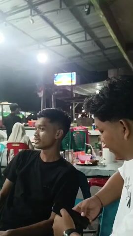 The video shows a group of people sitting at tables under a corrugated metal roof. The lighting appears to be from fluorescent bulbs overhead and a television screen in the background. In the foreground, two young men are visible. One is wearing a black t-shirt and looking off to the left, while the other, in a white t-shirt, is partially obscured and looking towards the right. In the middle ground, a woman in a black and white striped top and a headscarf is standing, and another person in a light-colored headscarf is seated on a red chair. Further back, two men are visible near a counter or food service area. The environment suggests an outdoor or semi-outdoor dining or gathering space, possibly a market or food stall area, with multiple tables and chairs set up.