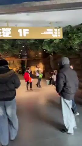 People walk down a wide, brightly lit hallway in what appears to be a museum or themed building. A large sign overhead reads "1F". The hallway is decorated with artificial foliage and stone-like structures that simulate a natural environment. Some people are wearing coats and hats, suggesting it is cool indoors. Two people in matching outfits walk in the foreground. There are several other people in the background, some standing, some sitting. The ceiling is lined with a modern-looking slat design.