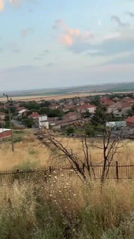 The video pans across a rural landscape at dusk. In the foreground, dry grass and a metal fence are visible. Beyond the fence, a village with red-tiled roofs nestles in a valley. Rolling hills, dotted with trees and power lines, form the background. The sky is filled with dramatic clouds, illuminated by the setting sun. A person's shoulder and upper back, wearing a black t-shirt with a purple flame design, are visible in the bottom right corner as the camera pans.
