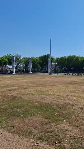 The video shows an outdoor park on a clear day. In the foreground, there is a dry, grassy field with some fallen leaves. In the middle ground, there are several park benches placed along a paved walkway. Behind the benches, there is a line of trees, and beyond the trees, a tall communication tower and a building with a traditional roof are visible. The sky is bright blue and cloudless. The camera pans slightly to the right.
