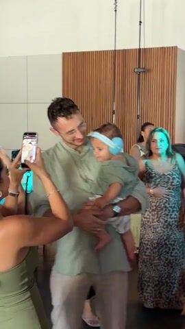 The video shows a man in a light green collared shirt holding a baby wearing light blue earmuffs. A person's arm with a bracelet and holding a smartphone is visible in the foreground on the left, filming the man and baby. The baby is looking down, and the man is smiling and looking at the baby. In the background, a woman in a leopard print dress is partially visible on the right. The environment appears to be indoors, with a wooden wall in the background. The lighting suggests an event or gathering.