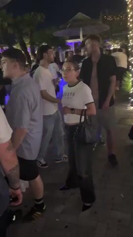 The video shows a crowded outdoor scene at night, possibly a bar or club, with many people gathered. There are lights, including string lights and some colored lights, illuminating the area. People are standing and walking around, some in groups. One person in the foreground is wearing a white t-shirt with "GUESS" on it, dark pants, and a black purse. Another person to their left is wearing a light blue collared shirt and dark shorts. The overall atmosphere appears to be lively and social, with a focus on people interacting and moving through the space.