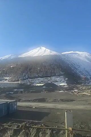 The video captures a daytime scene featuring a snow-covered mountain against a clear blue sky. At the base of the mountain, there's a heavily industrial area, including a gray building and piles of dark, granular material, resembling sand or gravel. A few blue containers are also visible on the left side of the frame. The mountain itself has trees growing along its slopes, and its peak is covered in a thick layer of snow. The overall view suggests a landscape where industrial activity exists in the vicinity of a mountainous, potentially cold, environment.