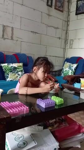 A young girl is sitting at a dark glass table in a living room. The girl is wearing a pink tank top and has dark hair pulled back. She is holding a small red spoon in her right hand and stirring something in a small, clear container in her left hand. On the table are several collections of large Lego-style blocks in pink, purple, and green. Behind the girl are two blue sofas with tropical leaf-patterned pillows. The walls are made of unfinished cinder blocks, and several framed pictures are hanging on the wall.  Under the table are a white box with a green umbrella symbol, a red plastic box, and other items. The girl appears focused on what she is mixing in the container.