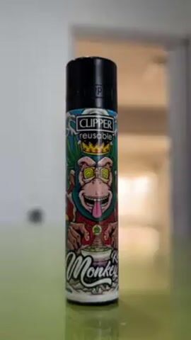 The camera zooms in on a reusable Clipper lighter. The lighter features a design of a cartoon monkey wearing a crown. The monkey has swirling eyes and is sticking its tongue out. The text "King Monkey" is written at the bottom of the lighter. The lighter is standing on a green surface. A blurred doorway is in the background.