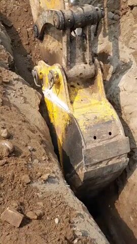 The video shows a close-up view of an excavator's yellow bucket digging a trench in the dirt. The bucket is partially submerged in the trench, scooping up soil. The trench walls are made of brown dirt and some small rocks. The excavator's arm and other machinery are visible above the bucket. The lighting suggests it is daytime.