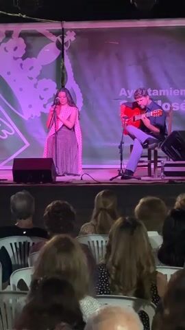 A live musical performance takes place on an outdoor stage. In the foreground, an audience sits in rows of white plastic chairs, facing the stage. The stage itself is illuminated with purple and pink lighting. On the stage, from left to right, are three performers and musical instruments. The performer on the far left is a woman wearing a dark, flowing dress, standing and singing into a microphone. Next to her, slightly to the right, is another woman, dressed in a patterned sleeveless dress, also singing into a microphone. On the far right, a man is seated on a stool, playing a red acoustic guitar. Behind the performers, a large green banner with a white floral pattern and text is displayed. The environment appears to be nighttime, with dark surroundings beyond the illuminated stage and audience area.